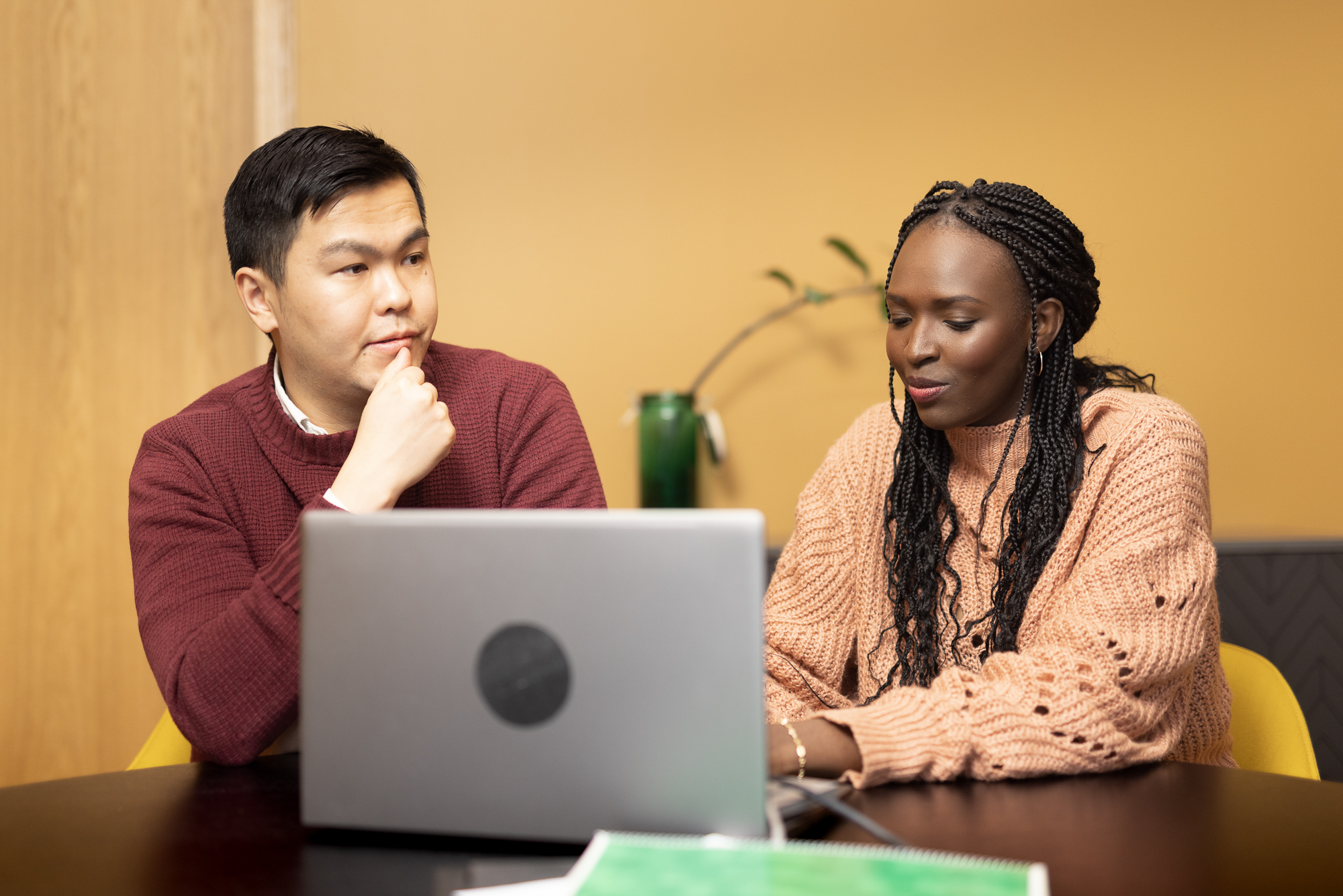 A woman and a man are sitting at a table. There is a notebook and a computer on the table.