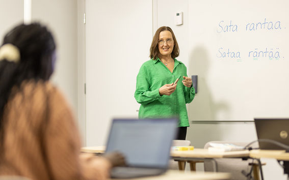 The teacher is standing in front of the class. There are two sentences in Finnish on the whiteboard: "Sata rantaa" and "Sataa räntää".