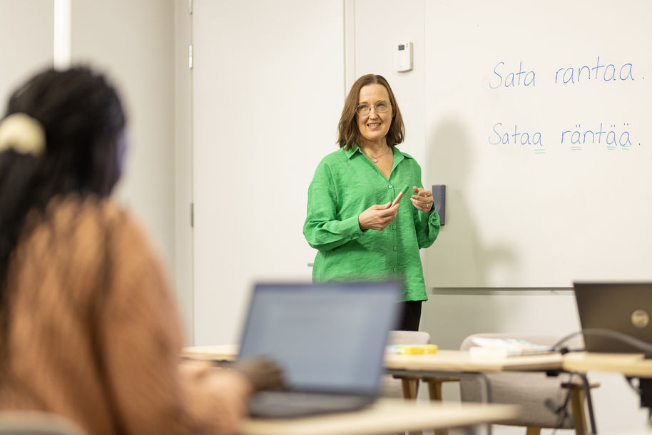 The teacher is standing in front of the class. There are two sentences in Finnish on the whiteboard: "Sata rantaa" and "Sataa räntää".